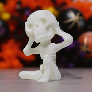 May include: A small, off-white skeleton figurine in a seated position, with its hands covering its ears. The figurine has detailed skeletal features and is set against a blurred background of Halloween decorations.