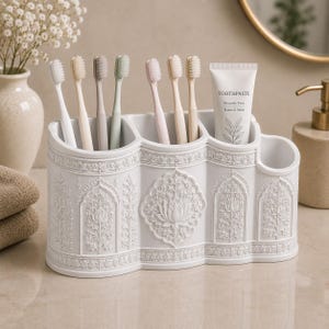May include: A white bathroom organizer with three compartments featuring intricate embossed floral and lotus patterns. It holds six toothbrushes in various pastel colors and a tube of toothpaste labeled Toothpaste Fluoride Free Natural Mint.