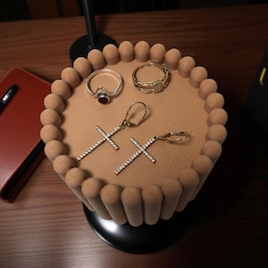 May include: A jewellery display featuring a silver ring with a red stone, a gold bracelet, and two pairs of earrings with cross pendants. The display is light brown and has a decorative edge.