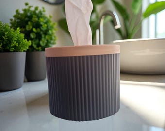 May include: A cylindrical tissue dispenser featuring a dark gray, vertically ribbed exterior body and a smooth, light pink circular lid. A single white tissue is pulled upward through the center opening of the lid, extending vertically.