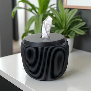 May include: A black tissue box with a ribbed design. The box has a round shape with a flat top and a slit for tissue dispensing. White tissues are pulled from the top. The tissue box is on a white surface with green plants in the background.