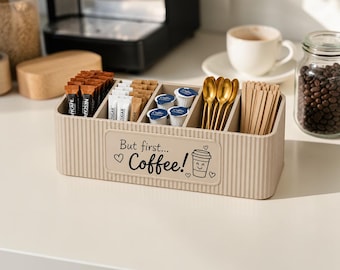 May include: A beige rectangular coffee station organiser with vertical ribbed sides. The front features the text "But first... Coffee!" with heart icons and a coffee cup graphic. It contains coffee packets, sugar sticks, creamer cups, gold spoons, and wooden stirrers.