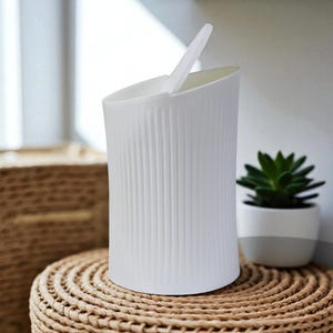 Può includere: Un cestino bianco con texture a coste e coperchio incernierato, posto su una superficie in vimini intrecciato. Una piccola pianta succulenta in vaso è nelle vicinanze. Il cestino ha un design moderno e minimalista.