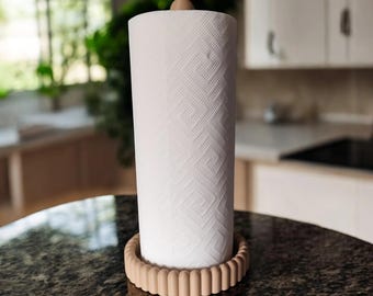 May include: A vertical paper towel roll with a white geometric embossed pattern, held by a light beige circular base with a fluted, scalloped edge and a matching light beige rounded finial topper that secures the roll in place.