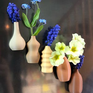 May include: Five small ceramic vases in different shapes and colours, each holding a different type of flower. The vases are arranged in a row on a dark surface. The flowers include blue forget-me-nots, blue hyacinths, and yellow primroses.
