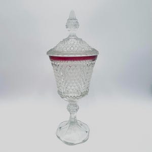 May include: A clear, stemmed glass candy dish with a diamond pattern and a lid. The rim of the dish features a band of deep red. The lid is topped with a pointed finial. The dish is elevated on a pedestal base.