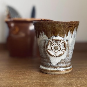 May include: A handcrafted ceramic mug with a brown and white glaze. The mug has a raised white floral emblem on the front. The mug is on a wooden surface, with a blurred brown object in the background.