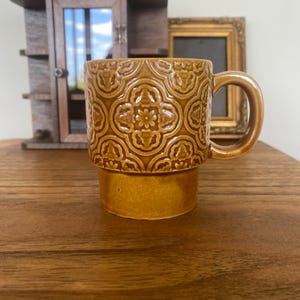 May include: A brown ceramic mug with a floral pattern. The mug has a rounded handle and a stacked base. The mug is on a wooden surface. A small wooden cabinet and a framed picture are in the background.
