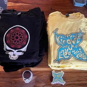 May include: Two folded t-shirts with graphic designs. The black t-shirt features a red and white geometric design over a white skull. The yellow t-shirt features a blue butterfly with the text "King Gizzard and the Lizard Wizard". A small sticker with a similar butterfly design is in the foreground.