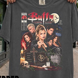 Könnte beinhalten: Ein anthrazitfarbenes T-Shirt mit einem Grafikdruck der Fernsehserie Buffy – Im Bann der Dämonen. Das Design zeigt den Titel, die Darsteller, einen Totenkopf, ein Kreuz, ein Buch mit Symbol und den Text Welcome to Sunnydale.