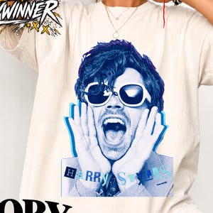 Puede incluir: Una camiseta de manga corta color marfil con un estampado gráfico de una persona con gafas de sol y las manos en la cara. El estampado es en tonos azules e incluye el texto "HARRY STYLES" en la parte inferior. Tiene cuello redondo.