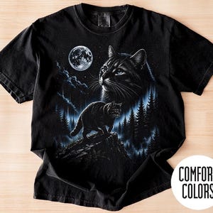 May include: A black short-sleeved t-shirt featuring a graphic print of a cat standing on a rocky cliff in a forest at night under a full moon. The design includes a large cat face in the sky above the forest. A Comfort Colors logo is visible.