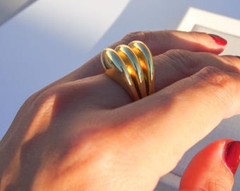 Può includere: Un anello dorato con un design unico e multistrato. L'anello è realizzato in metallo liscio e arrotondato e ha uno stile contemporaneo.