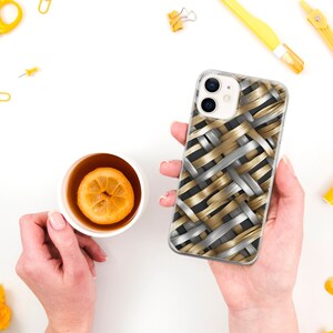 Puede incluir: Una funda transparente para tel&eacute;fono con un dise&ntilde;o tejido de bandas doradas y plateadas. La funda est&aacute; sujeta en una mano. Una taza de t&eacute; con una rodaja de lim&oacute;n est&aacute; en el fondo.