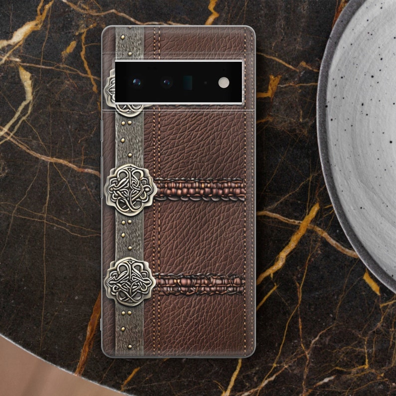 Puede incluir: Funda de tel&eacute;fono de cuero marr&oacute;n con superficie texturizada y detalles met&aacute;licos decorativos. La funda presenta una franja vertical con remaches y dos cierres circulares ornamentados. El dise&ntilde;o evoca una est&eacute;tica vintage.