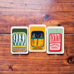 Può includere: Tre design di scatole di sardine. Il primo mostra sardine in una scatola verde con la parola "Sardines". Il secondo mostra sardine e un gatto nero in una scatola gialla. Il terzo mostra sardine in una scatola rossa con le parole "Sardines Wild Caught in Olive Oil".