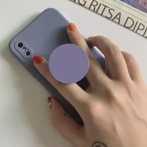 Puede incluir: Una funda de teléfono lila con un soporte para teléfono a juego. La funda del teléfono tiene una cámara y un flash. El soporte para teléfono es redondo y está adherido a la parte posterior del teléfono. El fondo es blanco con un periódico.