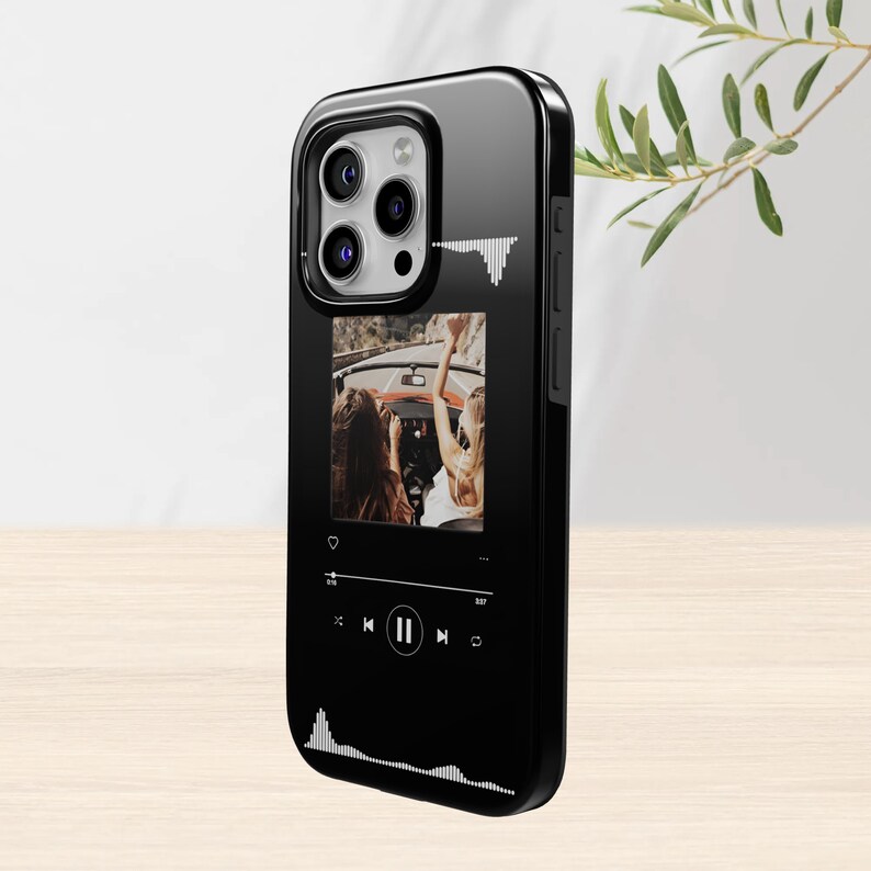 May include: Black phone case with a glossy finish. The case features a photo of two people in a car, with a music player interface design. White sound wave graphics are on the case.
