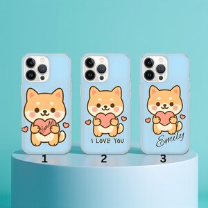 以下が含まれることがあります： 水色の携帯電話ケース3つ。それぞれ、漫画の柴犬のデザインが施されています。各ケースのデザインは異なり、「Emily」という名前が書かれたもの、「I LOVE YOU」と書かれたもの、そして「Emily」と書かれたものがあります。柴犬はハートを持っています。