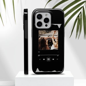 May include: Black phone case featuring a photo of two people in a convertible car. The case has a music player interface design with a play/pause button and a waveform graphic. The case is shown on a white surface.