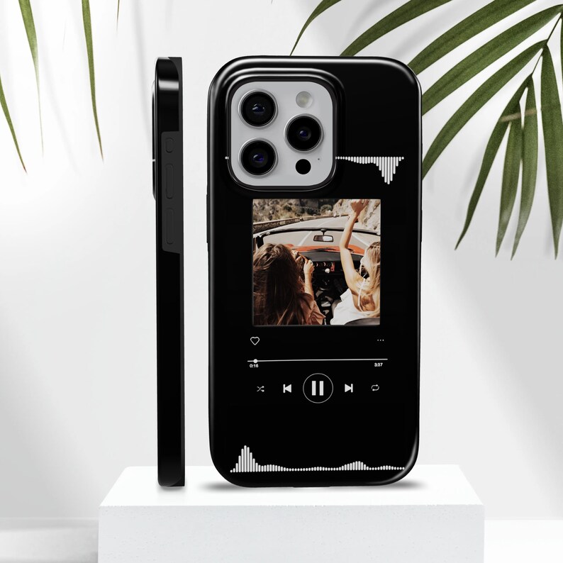 May include: Black phone case featuring a photo of two people in a convertible car. The case has a music player interface design with a play/pause button and a waveform graphic. The case is shown on a white surface.