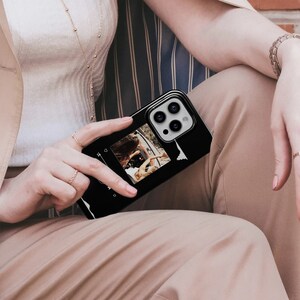 May include: Black smartphone case featuring a photo and a sound wave graphic. The case has a glossy finish and a raised camera module. The photo shows two people in a car. The case is held by a person wearing rings and a bracelet.