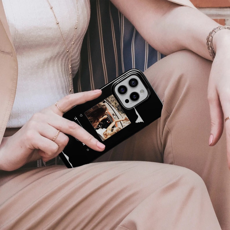 May include: Black smartphone case featuring a photo and a sound wave graphic. The case has a glossy finish and a raised camera module. The photo shows two people in a car. The case is held by a person wearing rings and a bracelet.