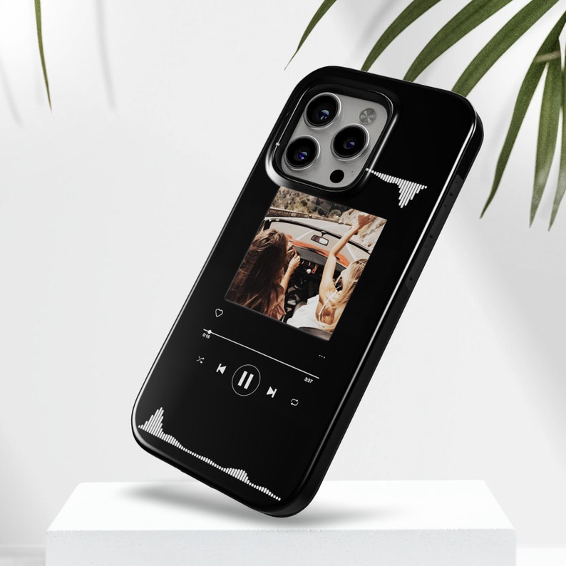 May include: Black phone case featuring a photo of two women in a convertible car. The case has a music player interface design with play, pause, and skip controls. White sound wave graphics are on the top and bottom.