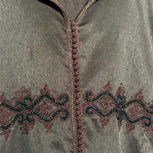 May include: A hooded, olive green garment with intricate embroidery in dark brown and forest green. The embroidery features a symmetrical design with floral and geometric elements. The garment has a central vertical line of decorative stitching.