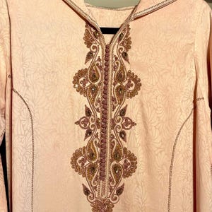 May include: A light peach-colored hooded garment with intricate gold and burgundy embroidery down the front. The fabric has a subtle floral pattern. The hood and edges are trimmed with gold detailing. The garment is hanging on a black hanger.