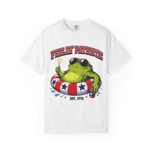 Pode incluir: Uma t-shirt branca de manga curta com o gráfico de um sapo verde com óculos de sol, a flutuar numa boia vermelha, branca e azul com padrão de estrelas. O sapo segura uma vela de faíscas e uma bebida. Texto: FEELIN' PATRIOTIC e EST. 1776.