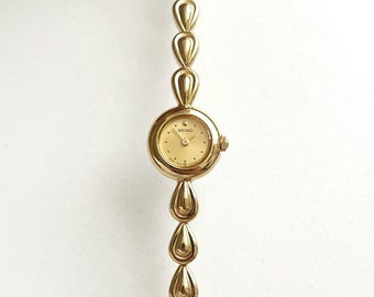 May include: A gold-tone Seiko wristwatch featuring a round analog dial with gold hands and markers. The watch band is composed of a series of polished, teardrop-shaped metal links that connect to form a continuous, fluid bracelet design.