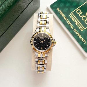 May include: A Gucci wristwatch featuring a two-tone silver and gold-tone metal link bracelet and a round case. The watch has a black textured dial with gold-tone hands, a date window at the six o'clock position, and the Gucci logo at the top.