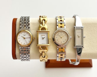 May include: Four elegant wristwatches displayed on a cream-colored stand. The watches feature silver and gold tones, with white watch faces. Brands include Longines, Gucci, and Hermès. The watches have various band styles, including link and bangle designs.