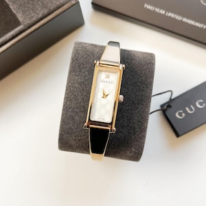 Puede incluir: Un reloj Gucci dorado con una esfera blanca rectangular con el logotipo de Gucci y un diseño estampado. El reloj tiene una correa dorada y un acento negro. El reloj se muestra sobre una superficie gris.
