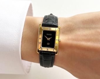 May include: A rectangular Gucci wristwatch featuring a gold-tone case with Roman numeral hour markers on the bezel, a black dial with gold hands, and a black leather strap with a textured crocodile-embossed finish and a gold-tone buckle.