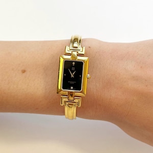 Puede incluir: Un reloj de pulsera dorado con una esfera negra rectangular. El reloj presenta una correa dorada y un pequeño acento de diamante. La esfera del reloj muestra la marca "Givenchy Paris".