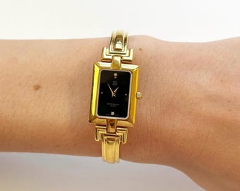 May include: A gold-toned wristwatch with a rectangular black face. The watch features a gold band and a small diamond accent. The watch face displays the brand name "Givenchy Paris".