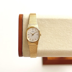 Può includere: Un orologio da polso Seiko Quartz color oro con un cinturino in metallo testurizzato a trama incrociata e una cassa rotonda. Il quadrante è di colore chiaro con sottili indici e lancette color oro, riportante il logo Seiko Quartz al centro.