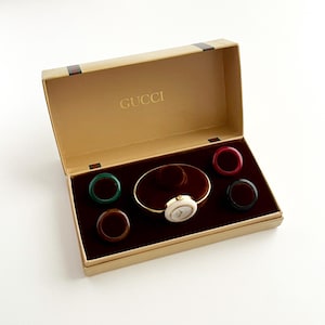 May include: A Gucci branded presentation box containing a gold-tone bangle watch with an interchangeable white bezel. The set includes four additional circular bezels in green, burgundy, black, and brown, all arranged on a dark velvet insert.
