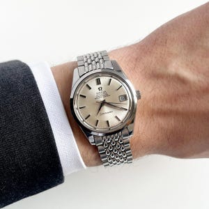 May include: A silver Omega Seamaster wristwatch with a light-colored face and a date display at the 3 o'clock position. The watch has a stainless steel bracelet and is worn on a wrist. The watch face displays the Omega logo and the word "Seamaster".