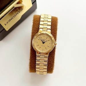 May include: A gold-toned Longines Quartz watch with a matching gold band. The watch face is a light gold color with black hands and markers. The watch is displayed on a brown velvet stand, with the Longines logo visible on the box.
