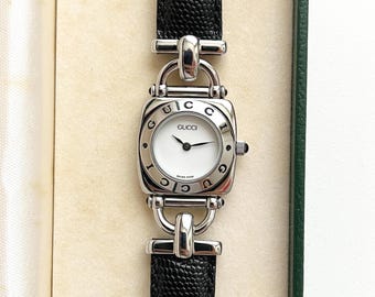 May include: A Gucci wristwatch featuring a white circular dial with black hands and the brand name printed in black. The silver-tone metal bezel is engraved with the word GUCCI repeatedly. It has a black textured leather strap with silver hardware.