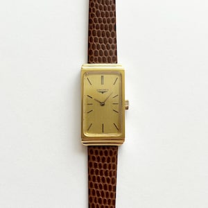 May include: A rectangular gold-tone Longines wristwatch featuring a gold dial with thin black hour markers and hands. The watch is attached to a brown leather strap with a textured, reptilian-inspired pattern and a small gold-tone winding crown.