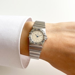 May include: A silver-toned Omega Constellation wristwatch featuring a round cream-colored dial with black Roman numeral hour markers on the bezel, a metallic link bracelet, and a crown on the right side, displaying the Omega logo and text.