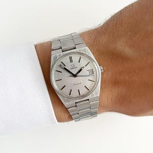 Relógio vintage raro OMEGA Genève 166.099 com mostrador prateado e data em aço.