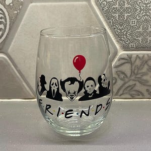 May include: A clear glass wine glass with a black and white silhouette of horror movie characters, including Pennywise, Ghostface, and Michael Myers. The text "FRIENDS" is printed below the characters with colorful dots.