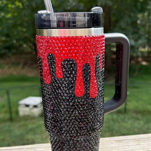 Puede incluir: Un vaso de acero inoxidable con asa y tapa negras. La parte superior es roja con un dise&ntilde;o de goteo, que pasa a negro en la parte inferior. Una pajita plateada est&aacute; insertada en la tapa. El vaso est&aacute; cubierto de brillantes pedrer&iacute;a.