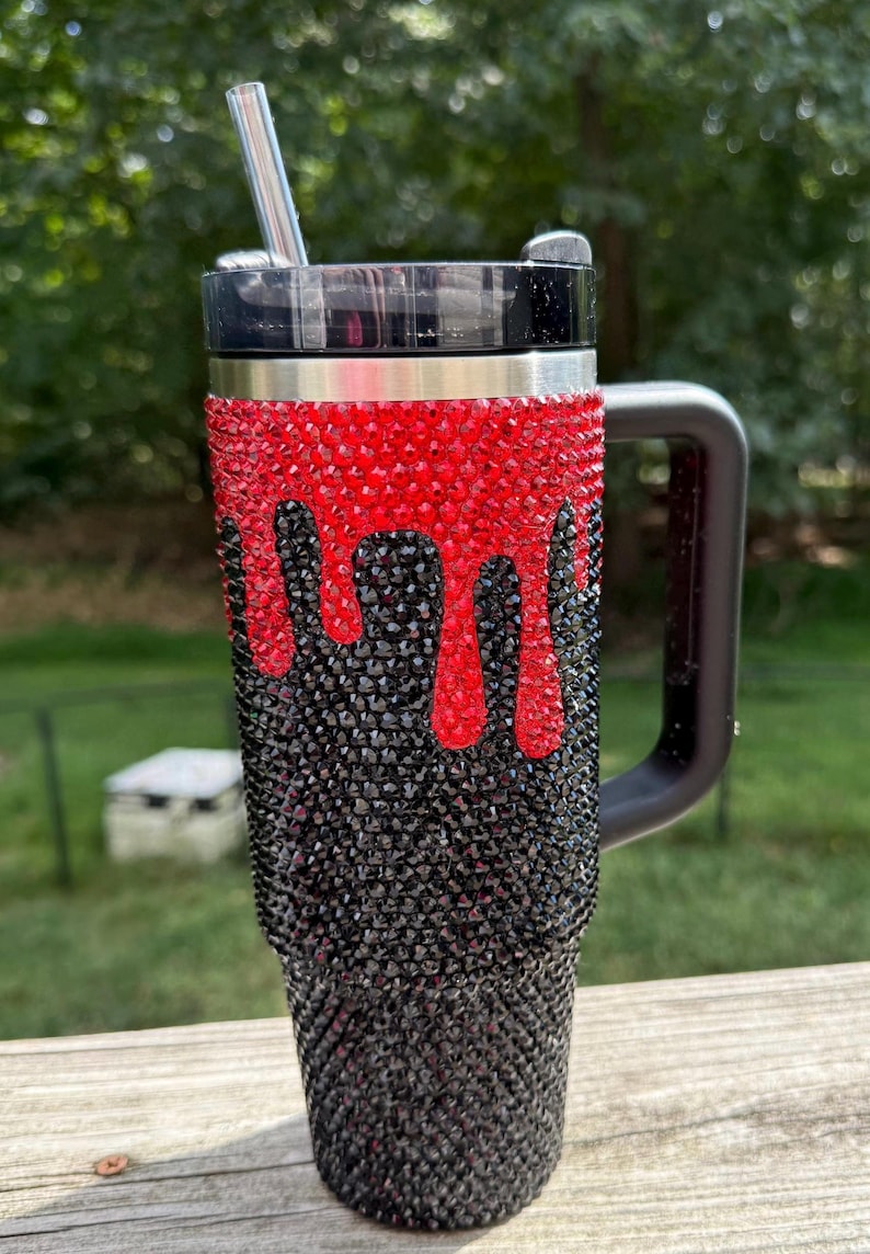 Puede incluir: Un vaso de acero inoxidable con asa y tapa negras. La parte superior es roja con un dise&ntilde;o de goteo, que pasa a negro en la parte inferior. Una pajita plateada est&aacute; insertada en la tapa. El vaso est&aacute; cubierto de brillantes pedrer&iacute;a.