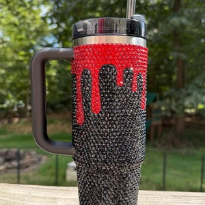 Puede incluir: Un vaso adornado con pedrer&iacute;a con asa y tapa negras. La secci&oacute;n superior est&aacute; cubierta de pedrer&iacute;a roja, con un dise&ntilde;o de goteo de pedrer&iacute;a negra que cae en cascada por el cuerpo. Una pajita plateada est&aacute; insertada en la tapa.